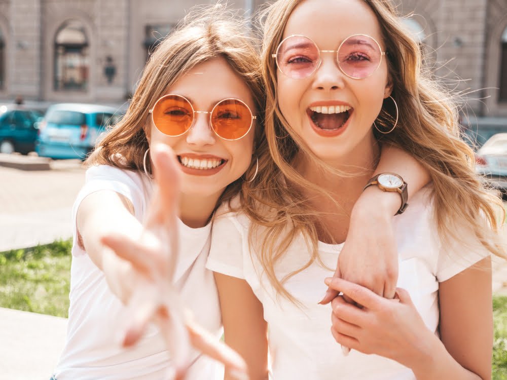 Two young women wearing stylish round sunglasses and smiling joyfully, with one reaching towards the camera, enjoying a bright day out together - Mirkin Vision