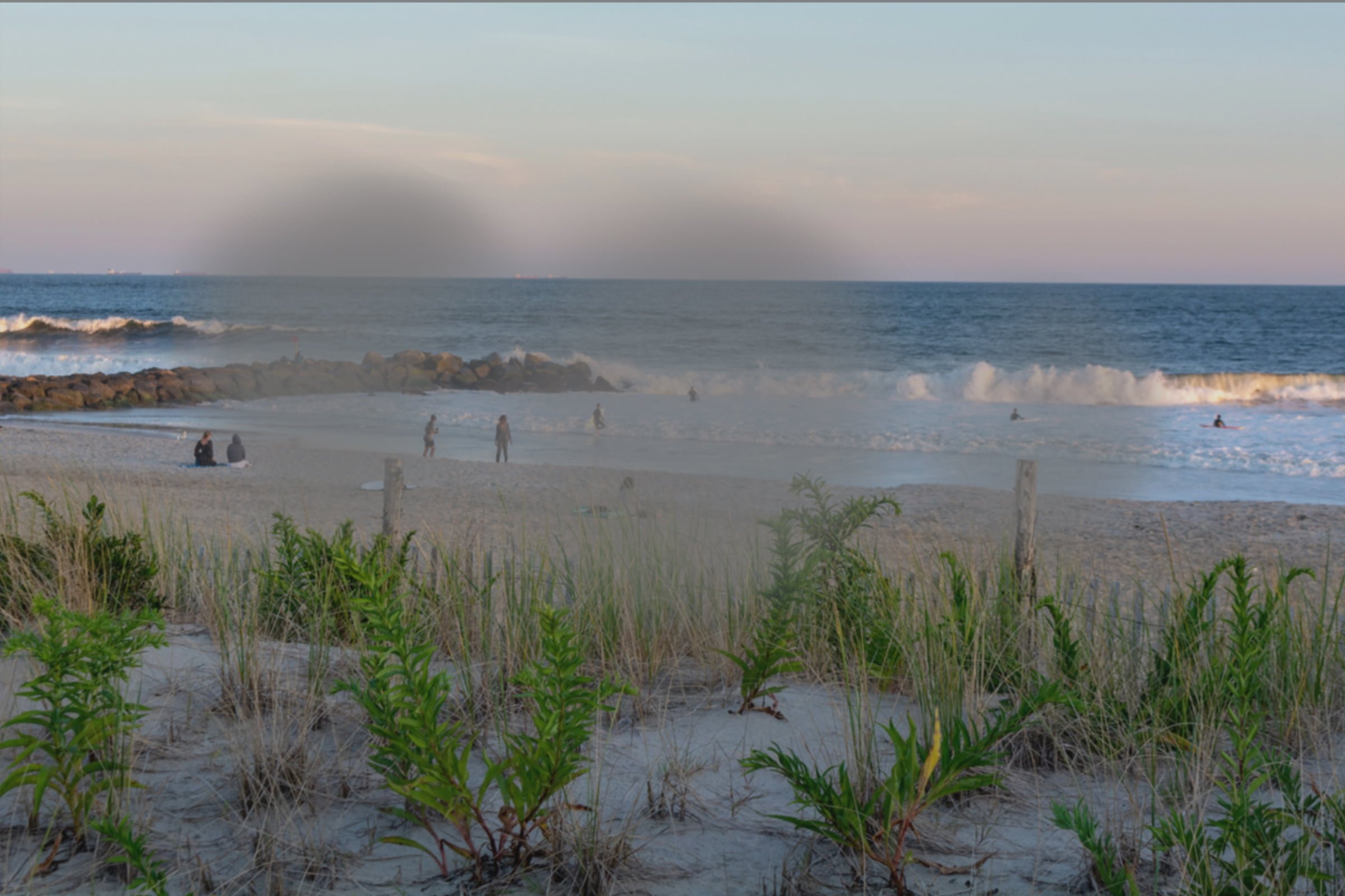 Age-Related Macular Degeneration vision on a serene beach scene with gentle waves, a few people on the sandy shore and in the water, and green dune grass in the foreground under a soft evening sky. - Mirkin Vision