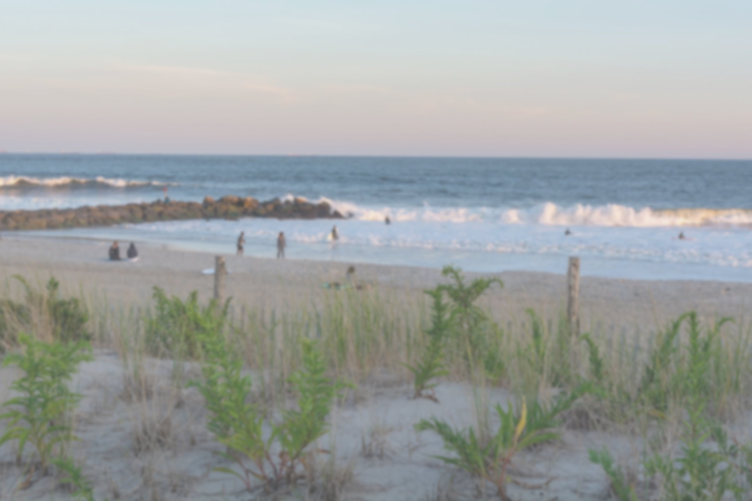 Cataract vision on a serene beach scene with gentle waves, a few people on the sandy shore and in the water, and green dune grass in the foreground under a soft evening sky. - Mirkin Vision