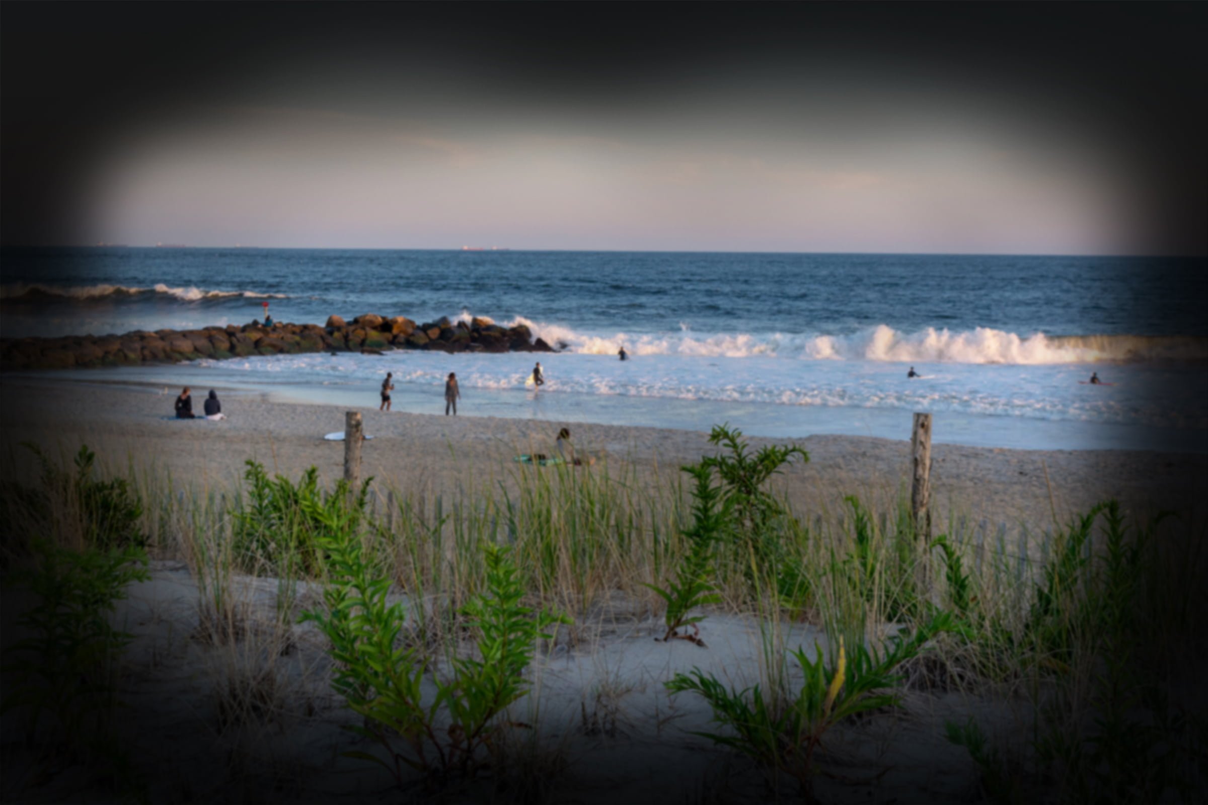 Glaucoma vision on a serene beach scene with gentle waves, a few people on the sandy shore and in the water, and green dune grass in the foreground under a soft evening sky. - Mirkin Vision