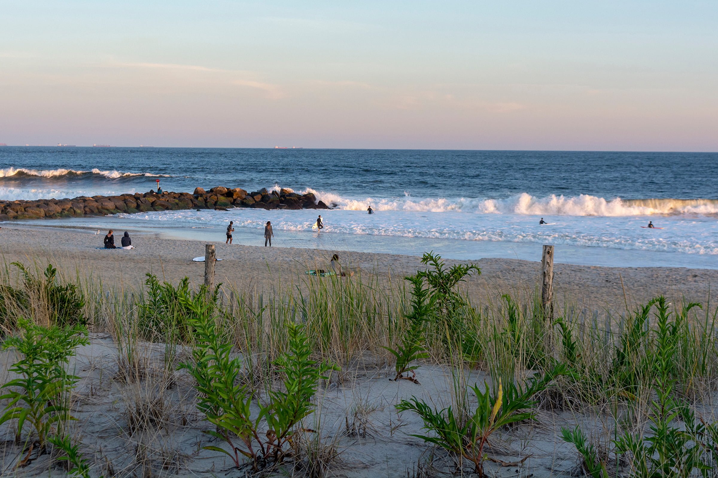 A serene beach scene with gentle waves, a few people on the sandy shore and in the water, and green dune grass in the foreground under a soft evening sky - Mirkin Vision