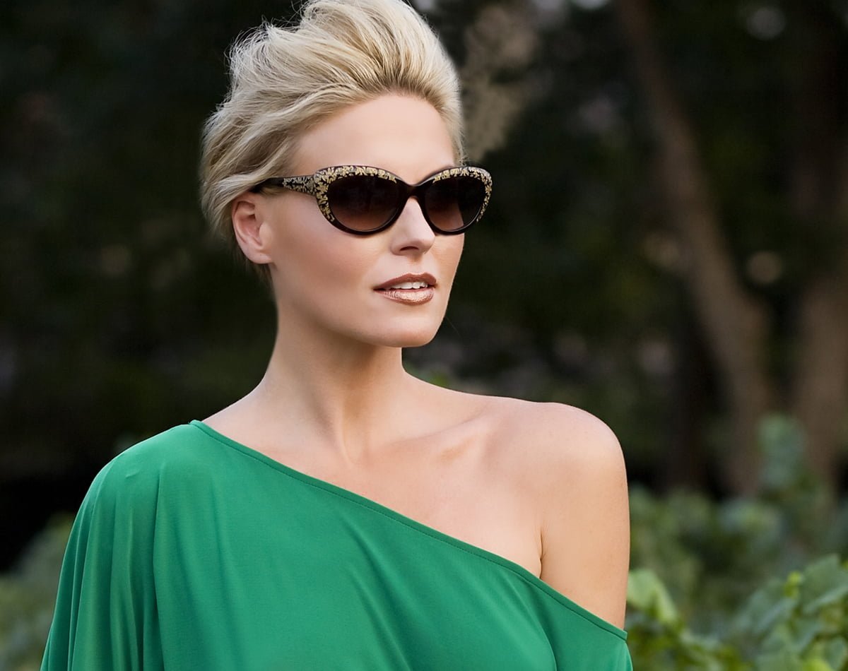  A stylish woman wearing oversized, patterned sunglasses and an off-shoulder green dress, with her hair swept back, standing outdoors against a blurred natural background - Mirkin Vision